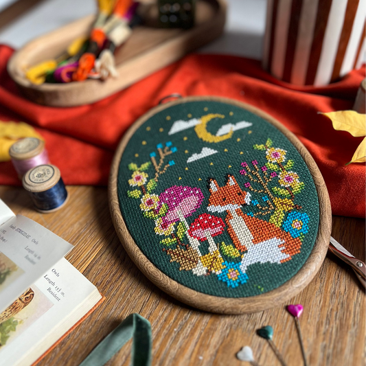 A finished cross-stitch embroidery piece in an oval wooden hoop, featuring a whimsical forest scene with a fox, mushrooms, flowers, and a crescent moon on a dark green background. The hoop is styled on a wooden table surrounded by craft supplies, including spools of thread, pins, ribbon, and a striped mug.