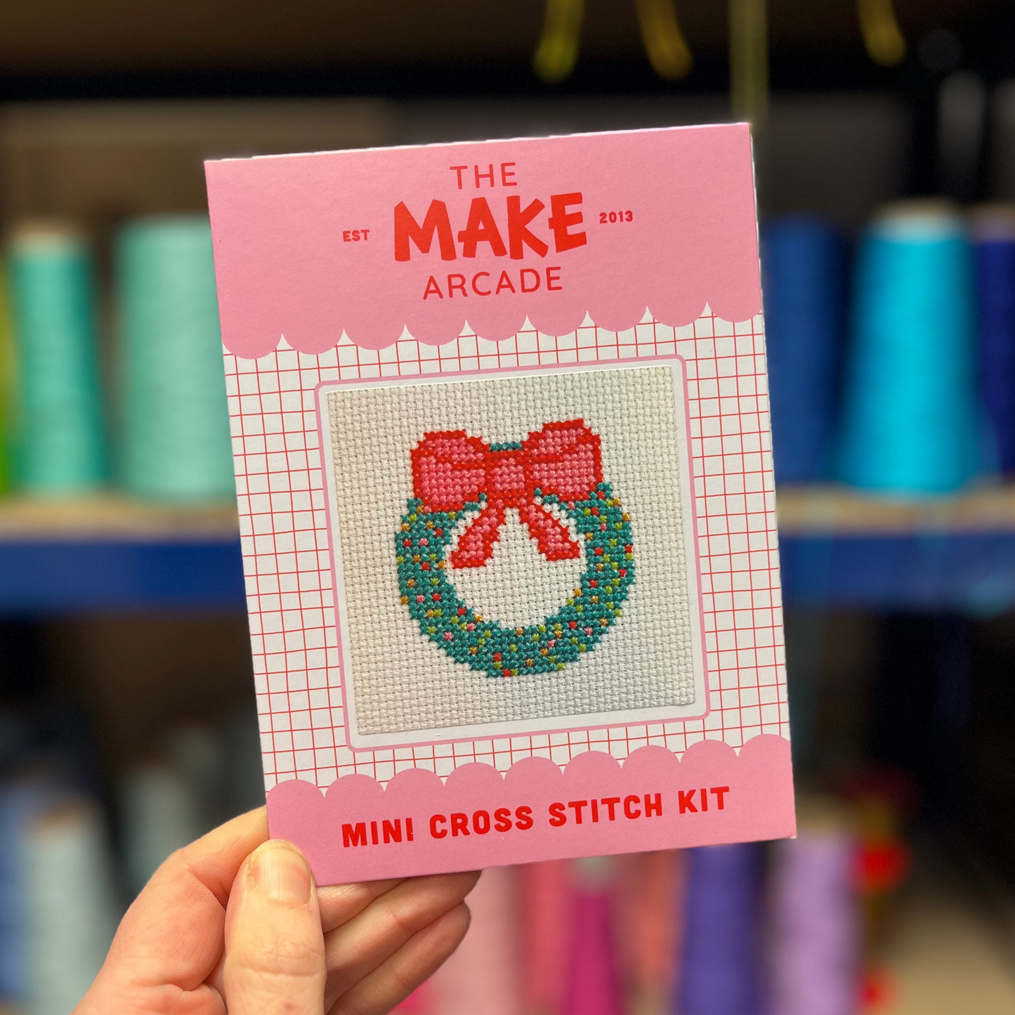 Make Arcade mini cross stitch kit with a wreath design held in front of a shelf with colorful items