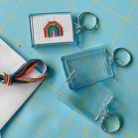 Cross-stitched rainbow design in a clear keyring frame on a blue cutting mat with keychains and embroidery floss.