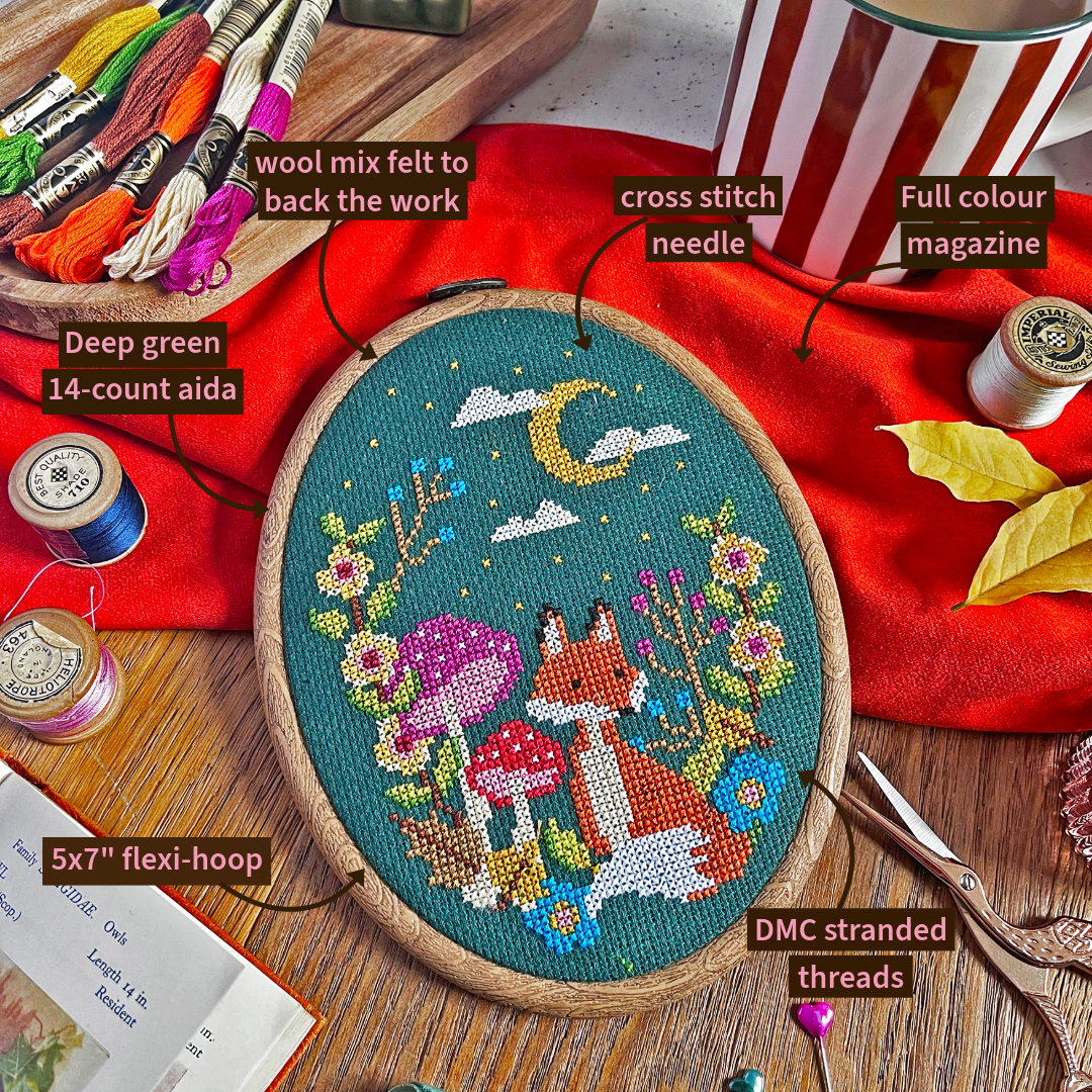 A completed cross stitch piece in a round wooden hoop, showing a whimsical woodland scene with a fox, mushrooms, flowers, and a crescent moon on a teal background. The hoop is photographed on a wooden surface with labeled callouts pointing to components such as wool mix felted to back the work, cross stitch needle, full colour magazine, deep green 14-count aida, 5x7" flexi-hoop, DMC stranded threads, and spools of thread.