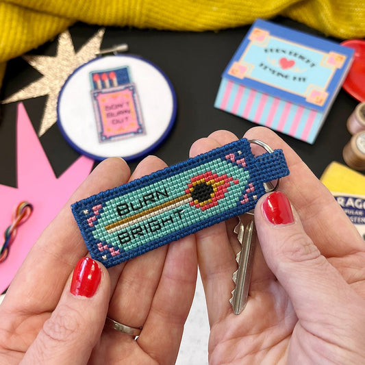 Hand holding a colorful embroidered key fob with 'Burn Bright' text, surrounded by other embroidered items on a table.