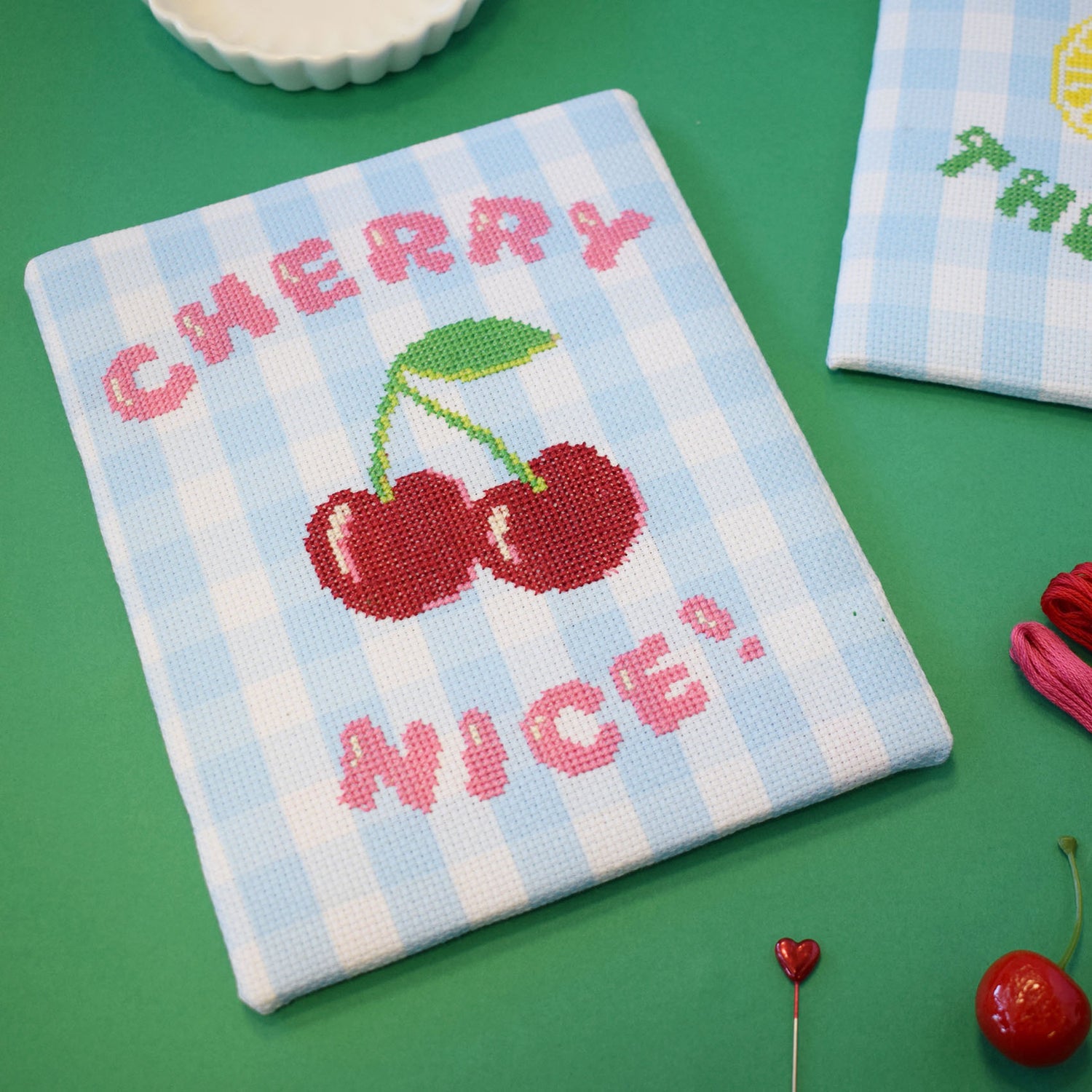 cross stitch design on blue gingham aida. The design is has the motif cherry nice with a large red cherry motif. The project is positioned on a deep green background.