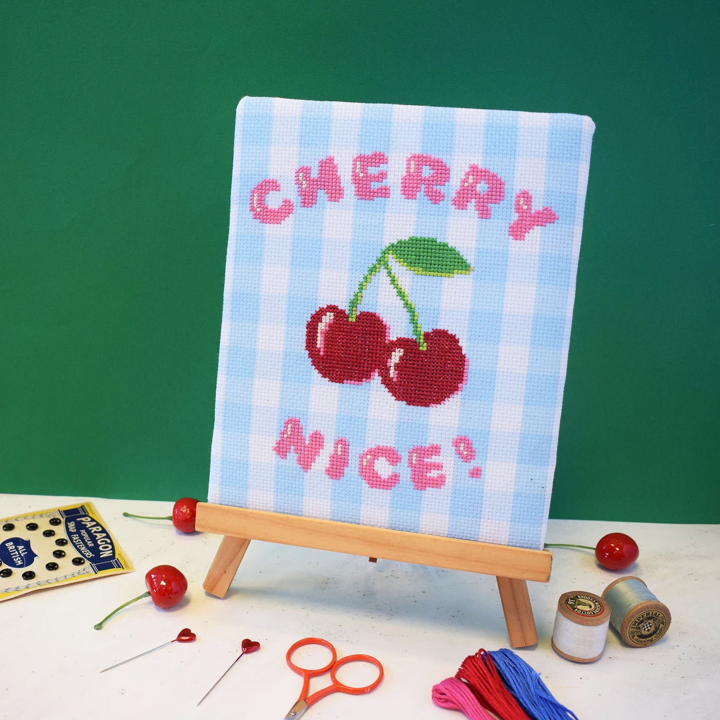 cross stitch design on blue gingham aida. The design is on a easel and has the slogan cherry nice with a large red cherry motif.