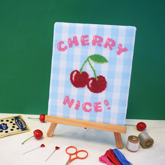 cross stitch design on blue gingham aida. The design is on a easel and has the slogan cherry nice with a large red cherry motif.