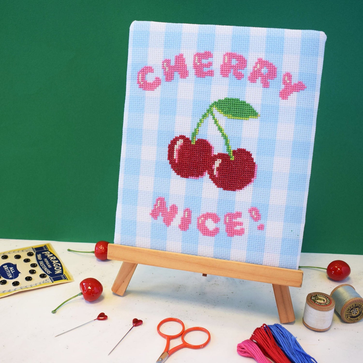 a cross stitch kit with cherry nice motto and cherries design. The aida fabric is gingham and the project is on a little easel.