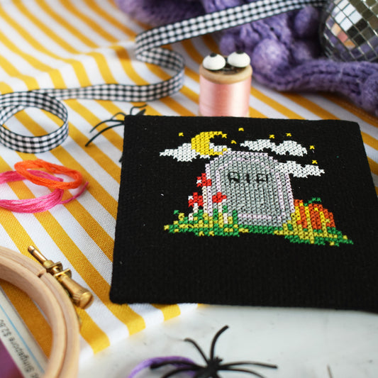 A small black embroidered canvas featuring a Halloween tombstone design with a crescent moon, clouds, flowers, and pumpkins, styled on a yellow and white striped fabric with embroidery supplies around it.