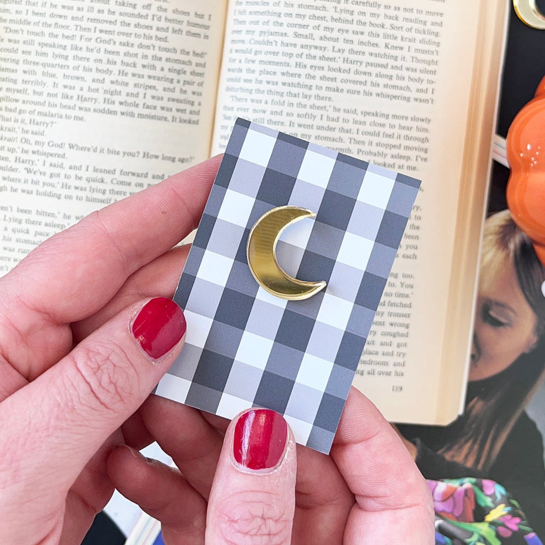 Hand holding a small checkered card with a gold crescent moon needle minder design over an open book.