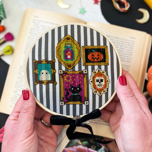 hands holding a hoop with Halloween-themed cross-stitch design with frames on a black and white stripe fabric