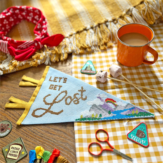 Cross-stitched pennant with 'Let's Get Lost' text on a checkered cloth with a mug and decorative patches.