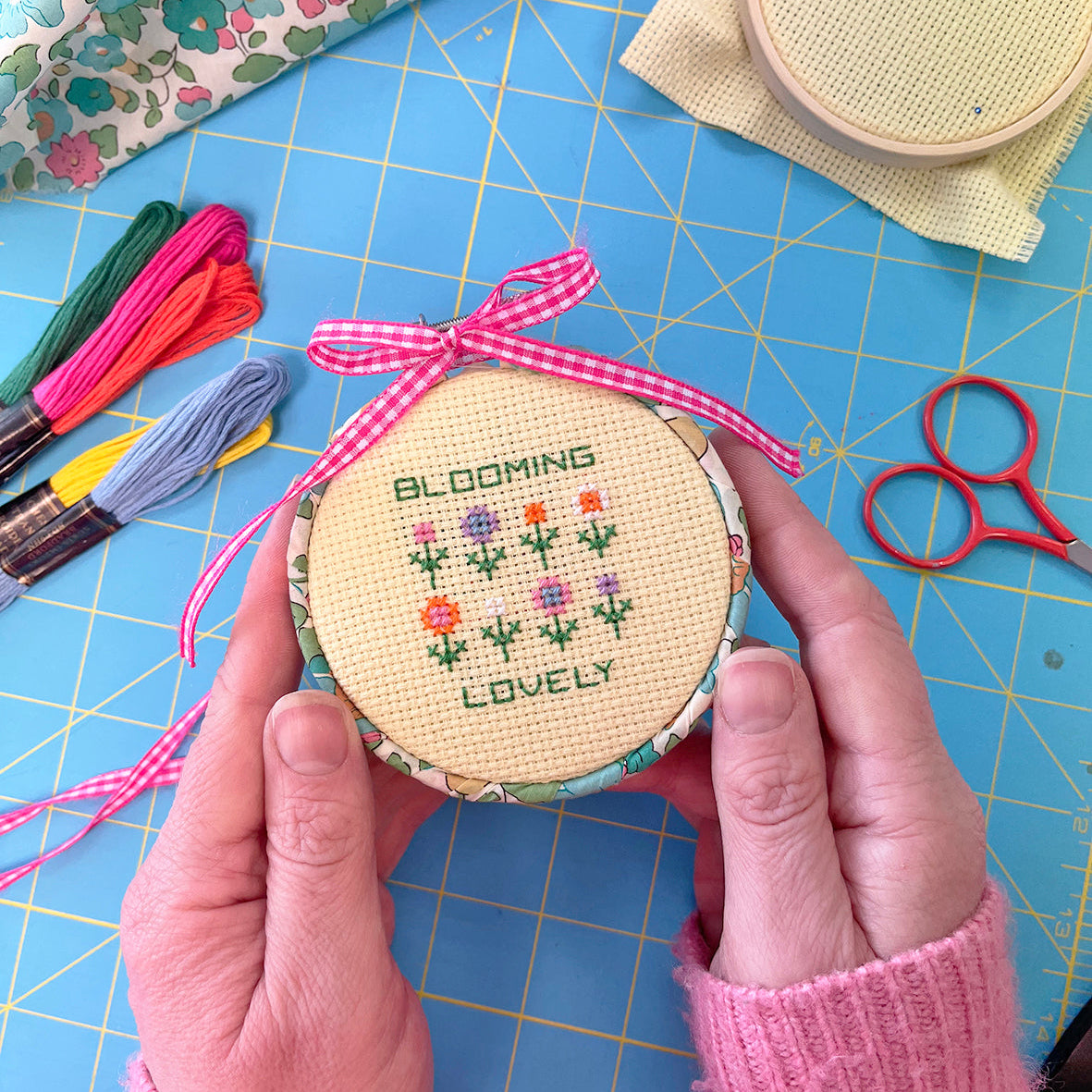 Hand holding a small cross stitch hoop with floral design on a blue cutting mat with sewing tools.