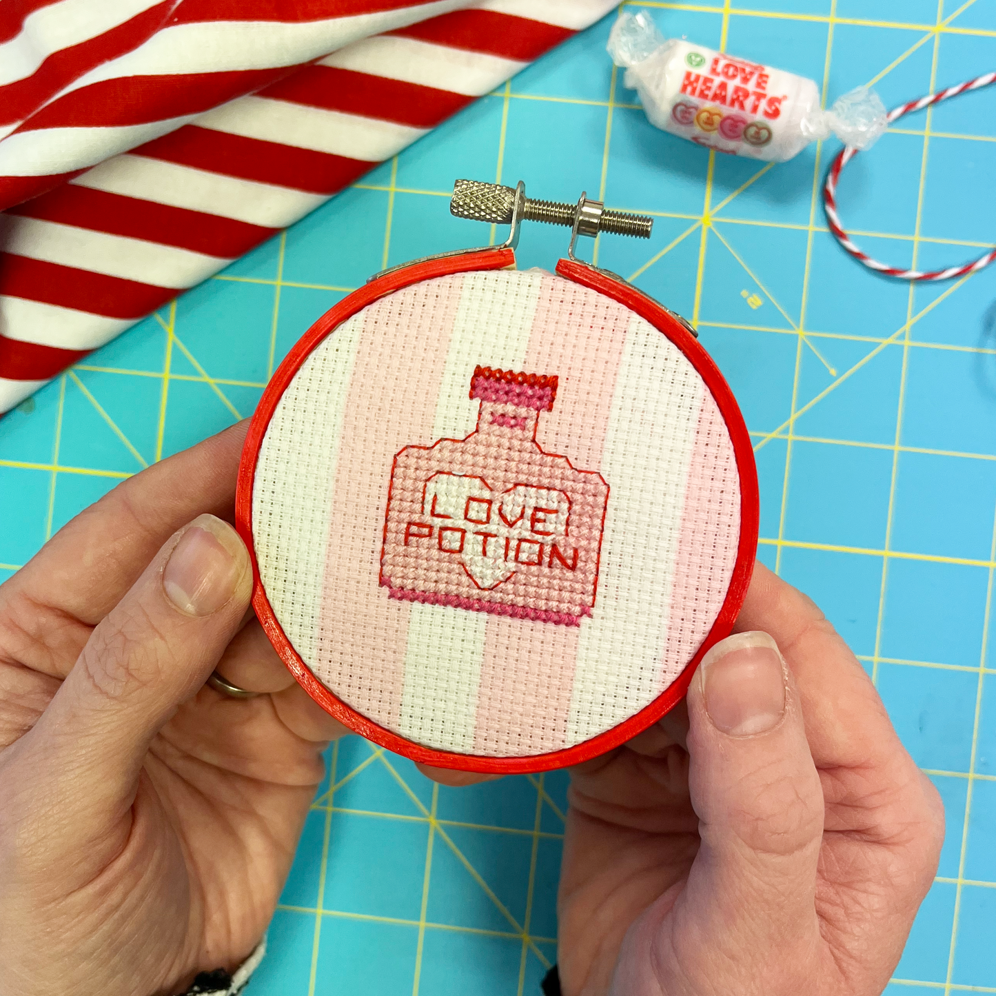 Hand holding a small embroidery hoop with 'Love Potion' cross stitch design on a blue cutting mat.