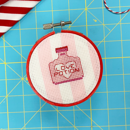 Embroidery hoop with 'Love Potion' cross stitch design on a blue cutting mat with candy and string.
