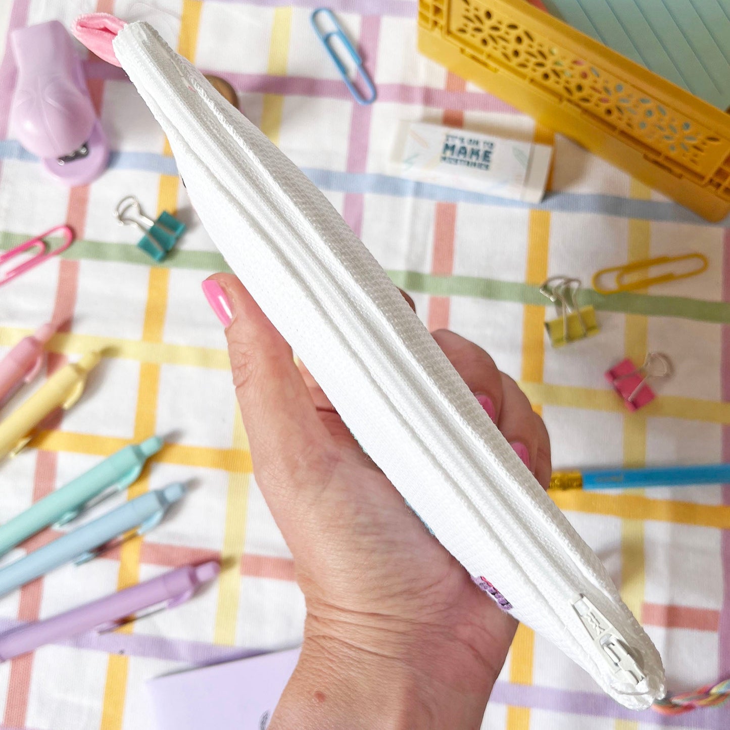 Hand holding a white cross stitch pencil case on a colourful checkered background with stationery items.