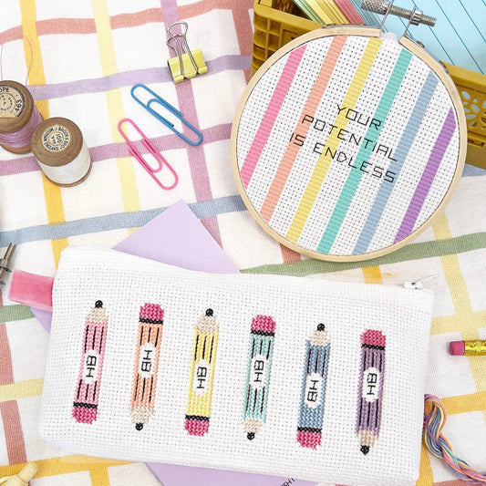 Stationery items including a cross stitch pencil case, pens, and an embroidery hoop with motivational text on a checkered background.