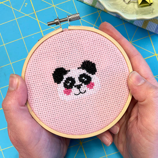 Cross-stitched panda design held in a wooden hoop on a blue cutting mat.