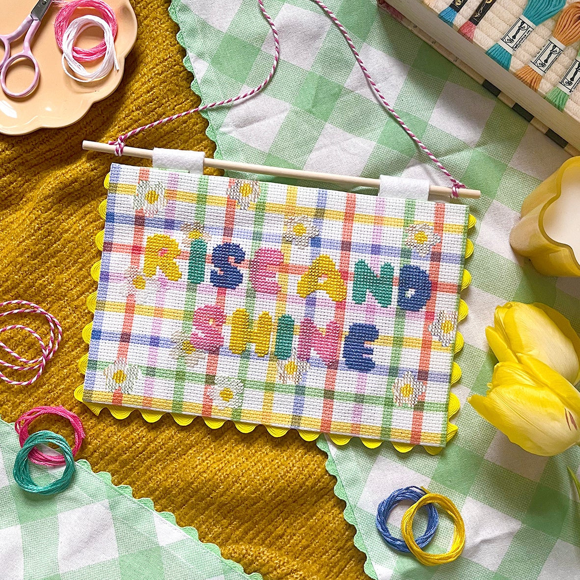 cross stitch sign with 'Rise and Shine' text on a checkered aida cloth with craft supplies.