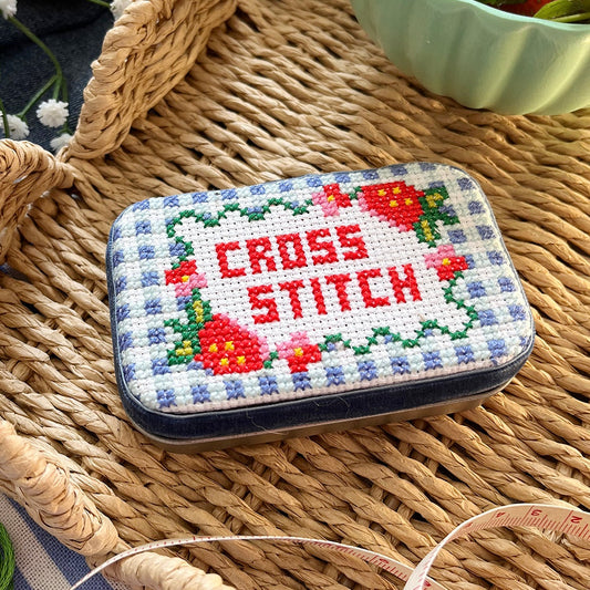 Cross-stitched tin with 'Cross Stitch' text on a woven surface with strawberries in the background