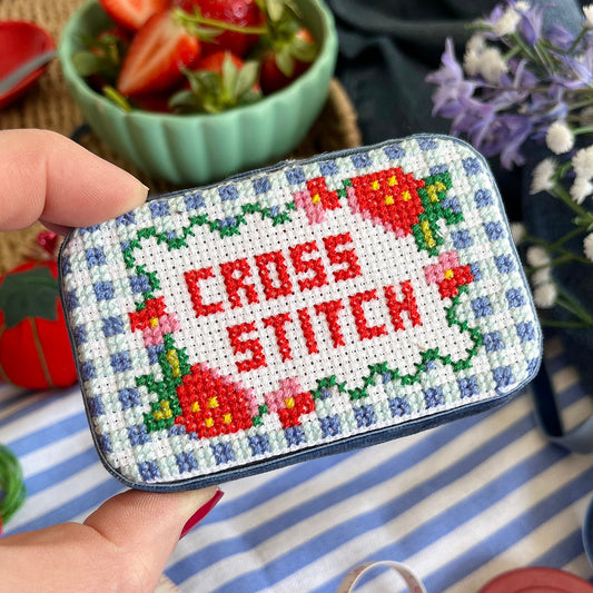 Cross-stitched tin with 'Cross Stitch' text held by a hand on a striped fabric background.