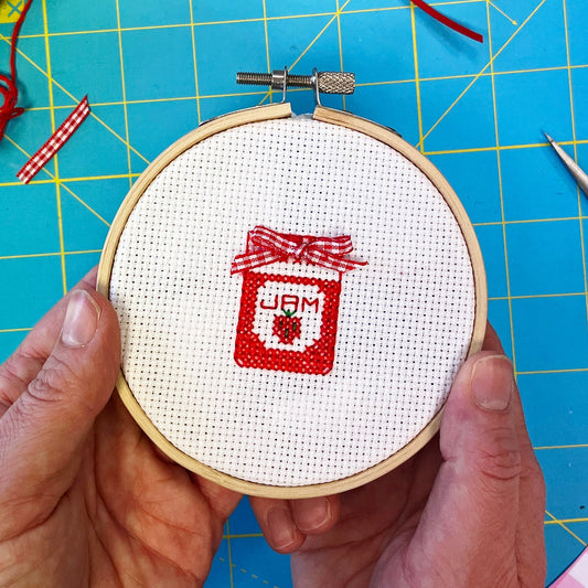 Cross-stitched design of a jar with 'Jam' text in a hoop on a blue cutting mat.