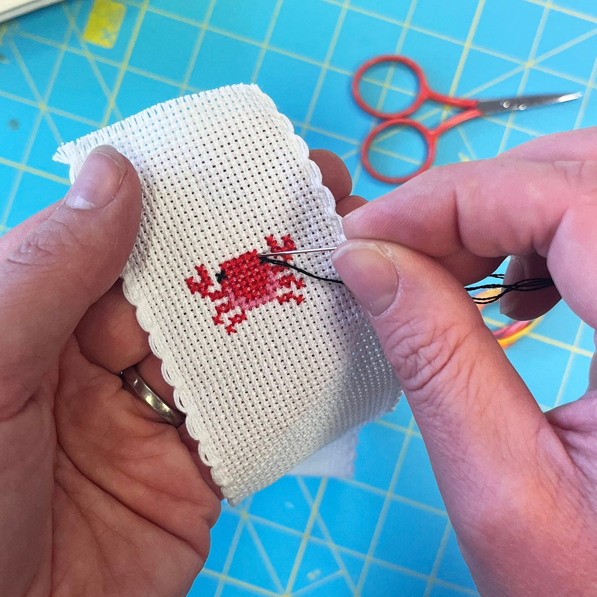 Person cross-stitching a crab design on a piece of bookmark fabric with red thread