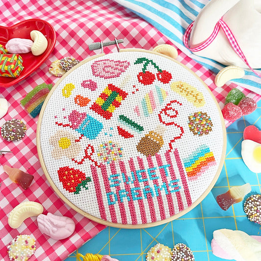 A round embroidery hoop displays a colorful cross-stitch design of sweets and the words 'SWEET DREAM' on white fabric. The hoop is surrounded by craft supplies, including spools of embroidery floss, candy pieces, a swan figurine, and striped fabrics on a bright blue grid surface.