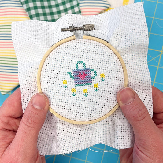 Embroidery hoop with watering can and flowers cross-stitched design held by hands on a colorful background