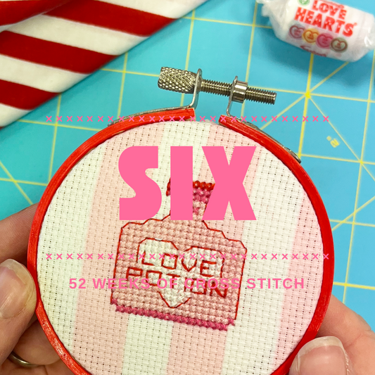 Embroidery hoop with a 'Love Potion' cross stitch motif on top of a blue cutting mat.