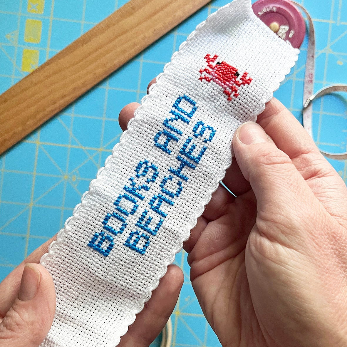 A person's hands holding a white cross-stitch sampler with blue embroidered text reading 'BOOKS AND BEACHES' and a small red crab motif. The background shows a blue cutting mat with a wooden ruler and sewing tools.