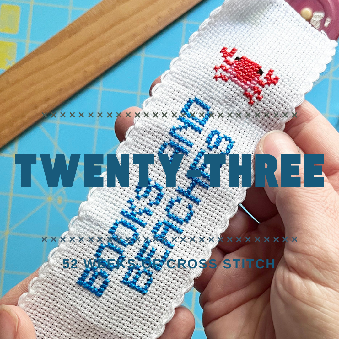 Hands holding a white cross-stitch sampler with blue cross stitch bookmark reading 'BOOKS AND BEACHES' and a red crab motif, over a blue grid cutting mat with a wooden ruler.