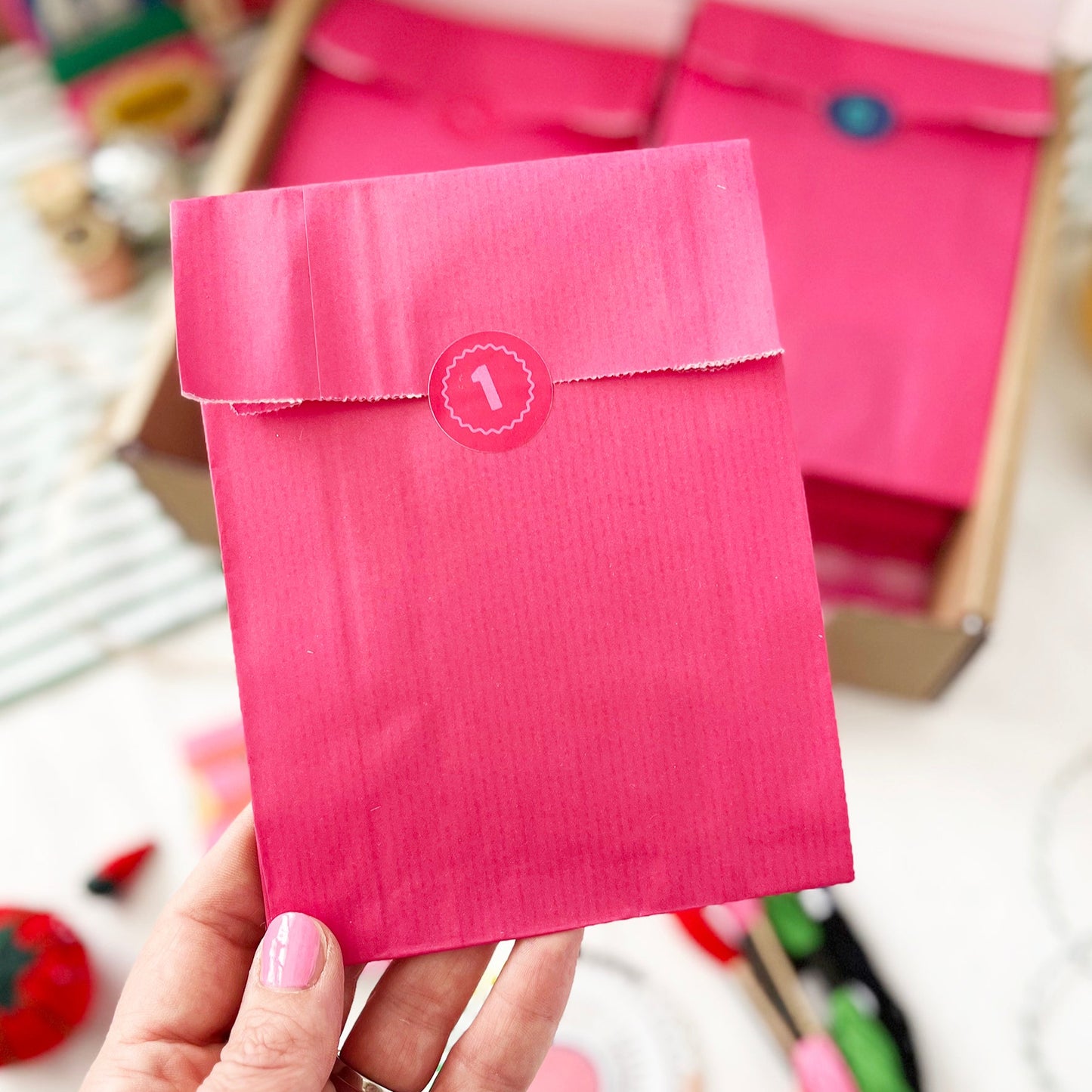 Hand holding a pink envelope from an advent calendar with a blurred background of similar envelopes and stationery items.