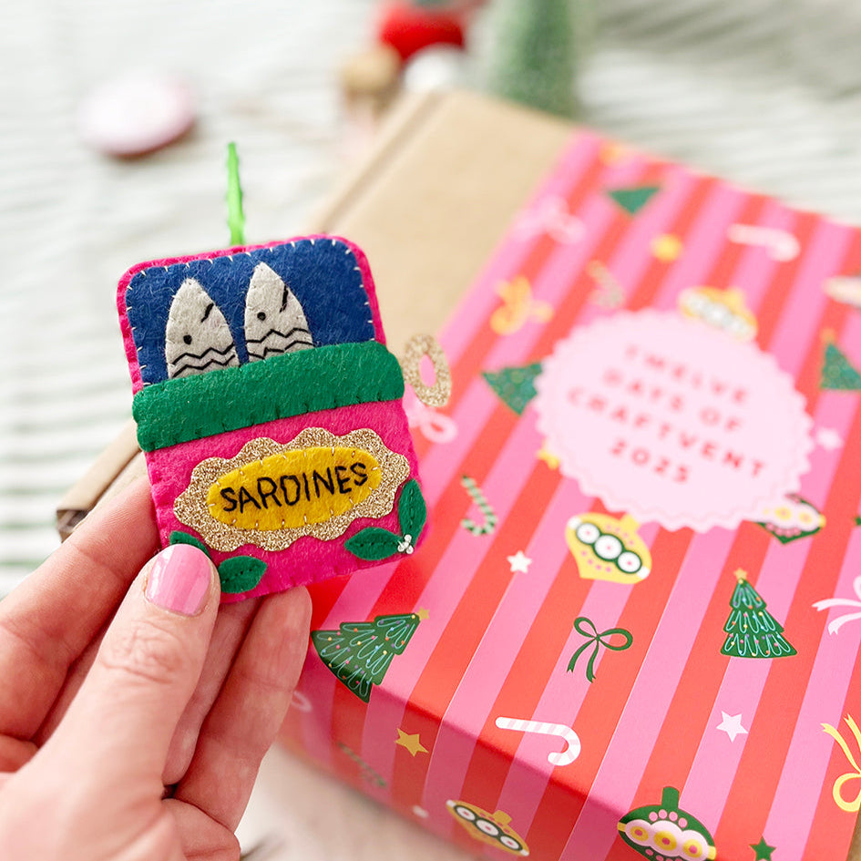 Hand holding a decorative christmas ornament in a sardine design on a pink striped box.