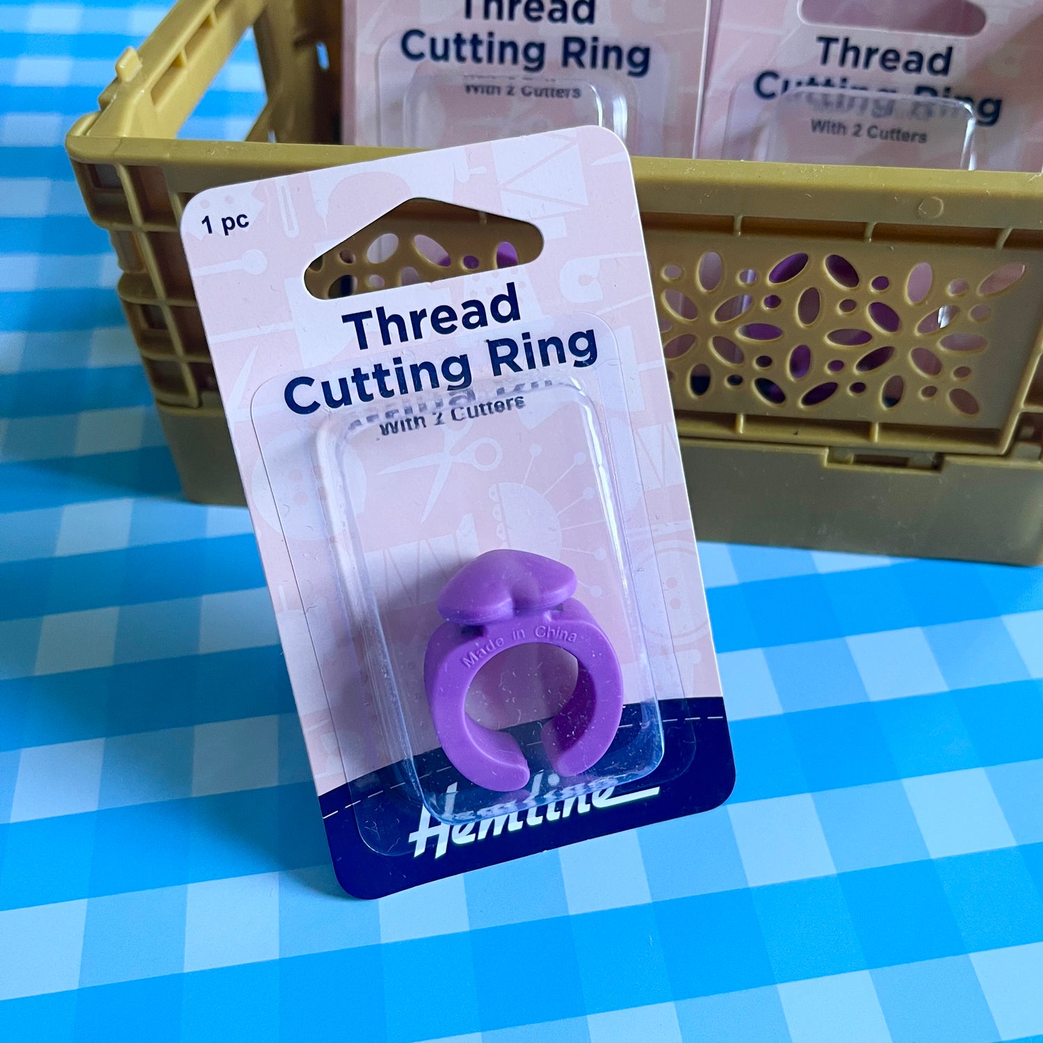 A purple thread cutting ring with two cutters, packaged on a white and blue Hemline card, placed in front of a tan plastic crate on a blue and white checkered surface.