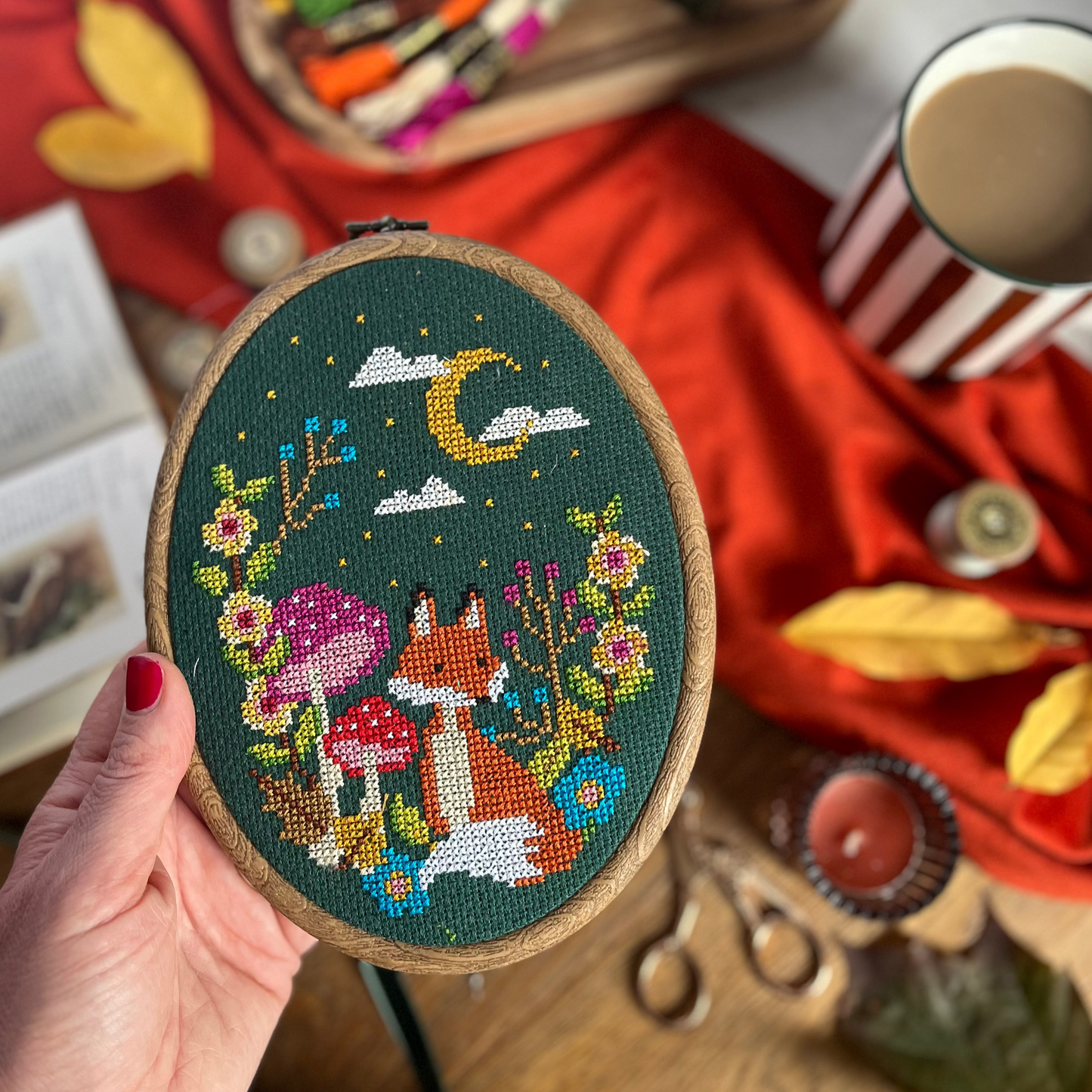 A hand holds an oval embroidery hoop featuring a colorful cross-stitch design of a fox, mushrooms, flowers, and a crescent moon on a dark green background. The scene is styled with a red fabric backdrop, autumn leaves, scissors, thread, and a striped mug.