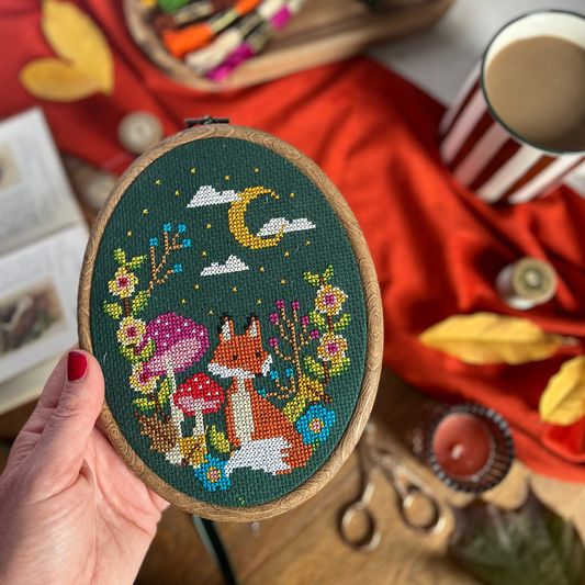 A hand holds an oval embroidery hoop featuring a colorful cross-stitch design of a fox, mushrooms, flowers, and a crescent moon on a dark green background. The scene is styled with a red fabric backdrop, autumn leaves, scissors, thread, and a striped mug.