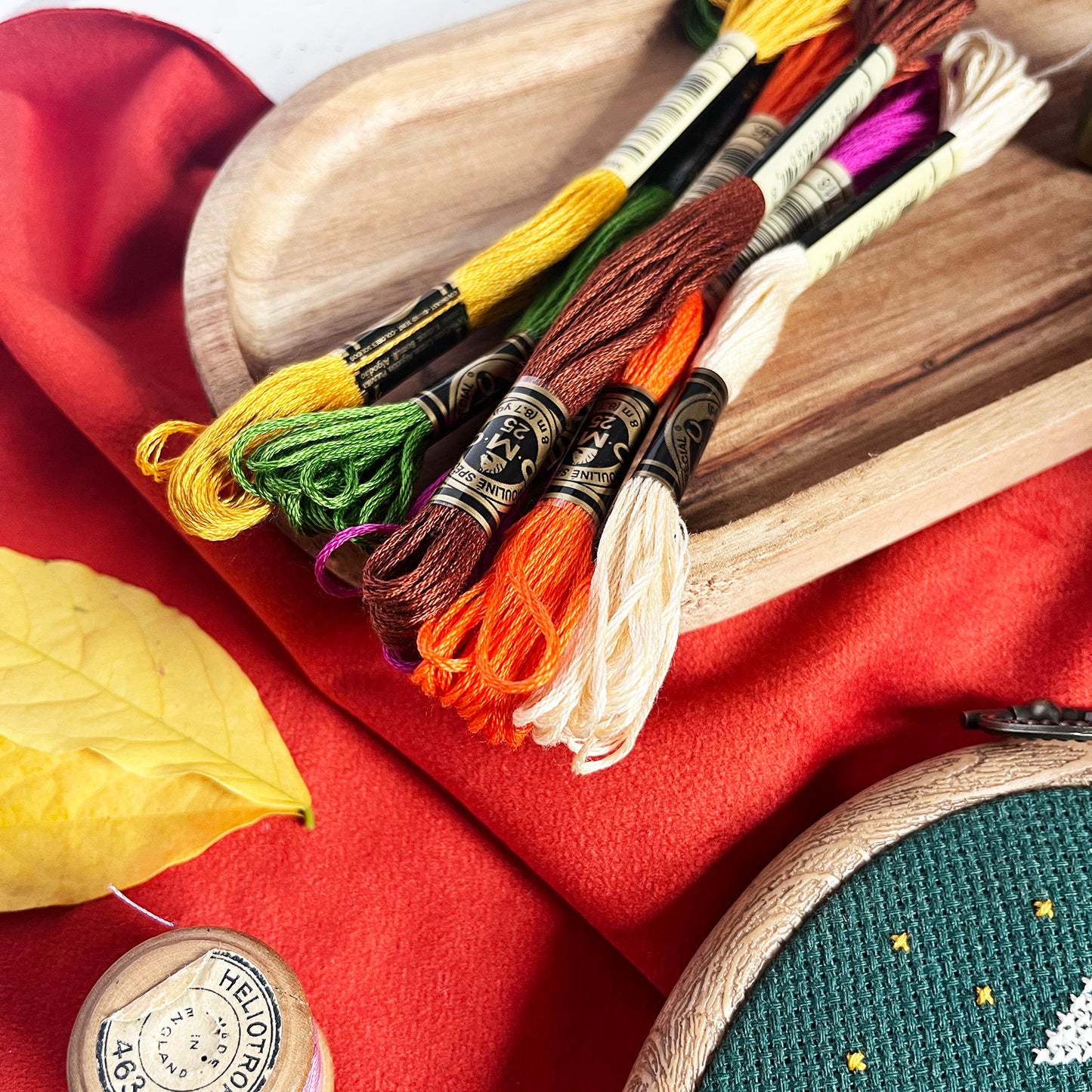 A set of embroidery floss thread skeins in assorted colors, including yellow, green, brown, orange, cream, and purple, arranged on a wooden tray over a red fabric background.