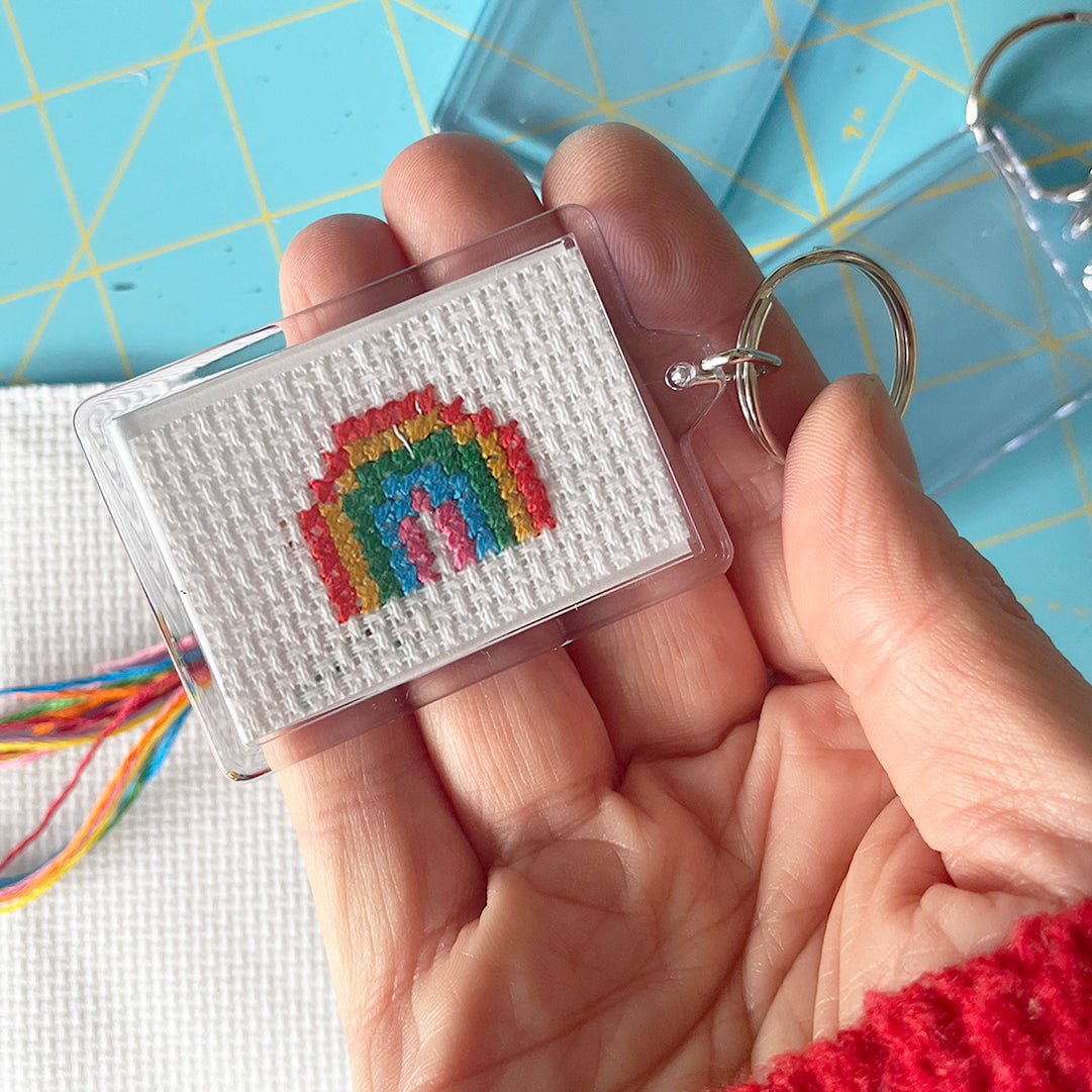 Hand holding a keychain with a rainbow cross-stitch design on a blue cutting mat.