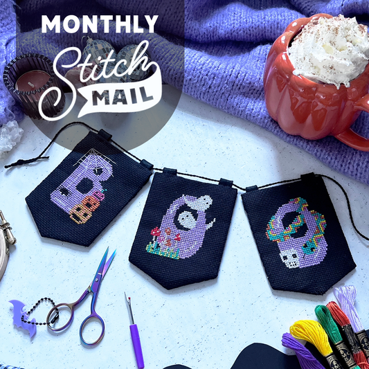 Three small cross-stitch flags are arranged on a light surface with craft tools and a hot chocolate. It is a halloween theme.