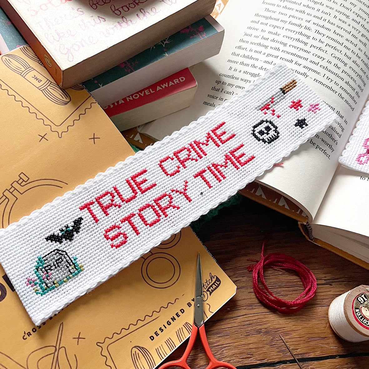 Cross-stitched bookmark with 'True Crime Story Time' text on a wooden surface with books and scissors.
