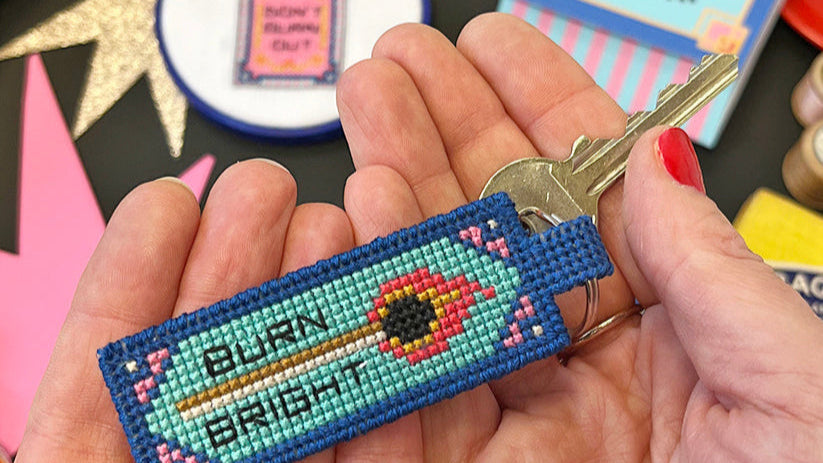 Keychain with colourful cross stitch design held between two hands against a colourful background.