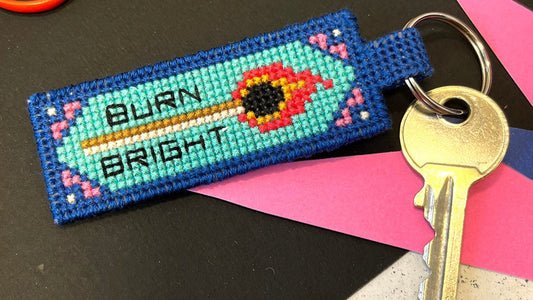 Cross-stitched key fob with 'Burn Bright' text on a black surface with crafting supplies.