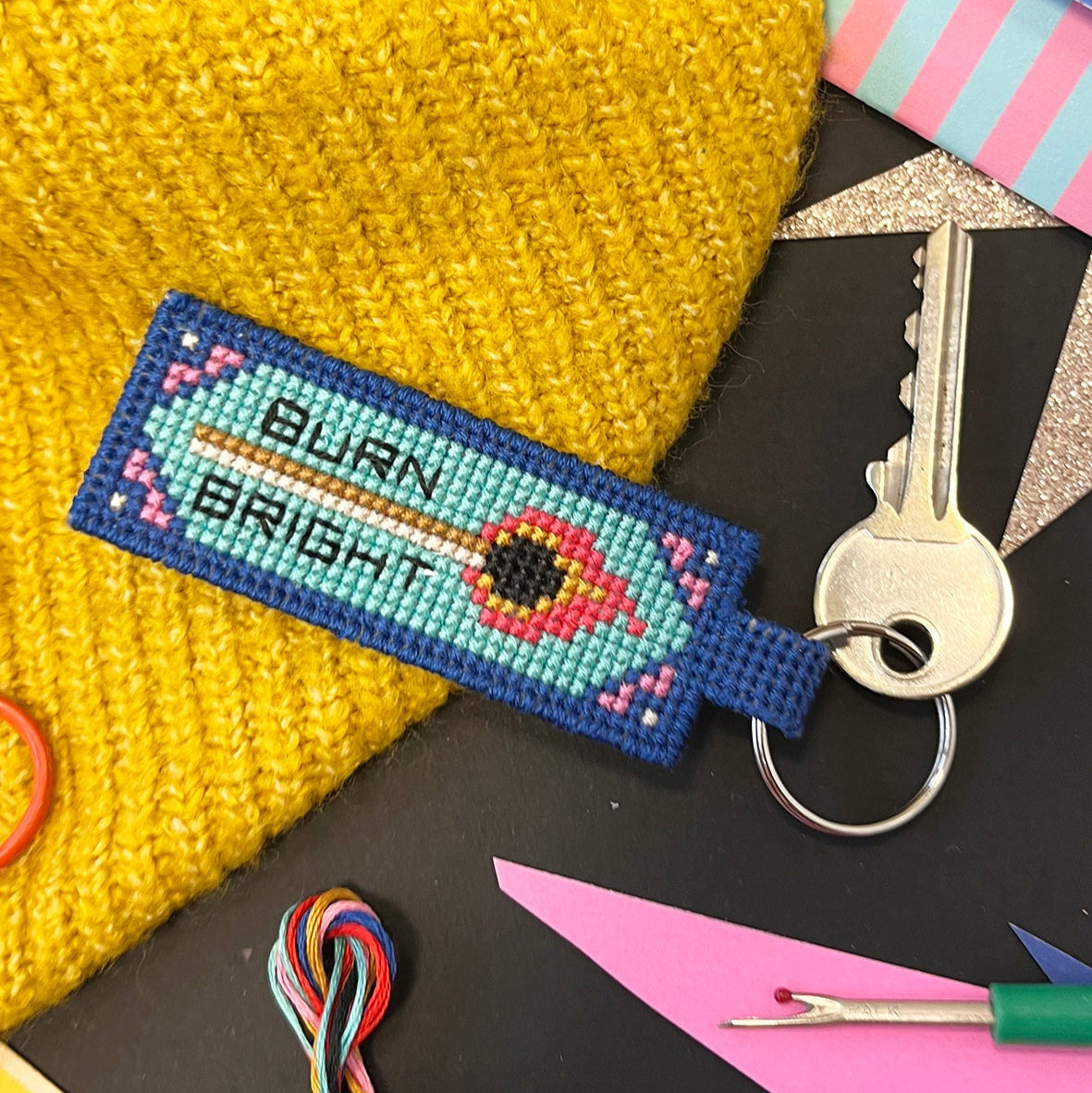 Cross-stitched key fob with 'Burn Bright' design on a black surface with a yellow beanie and colorful thread.