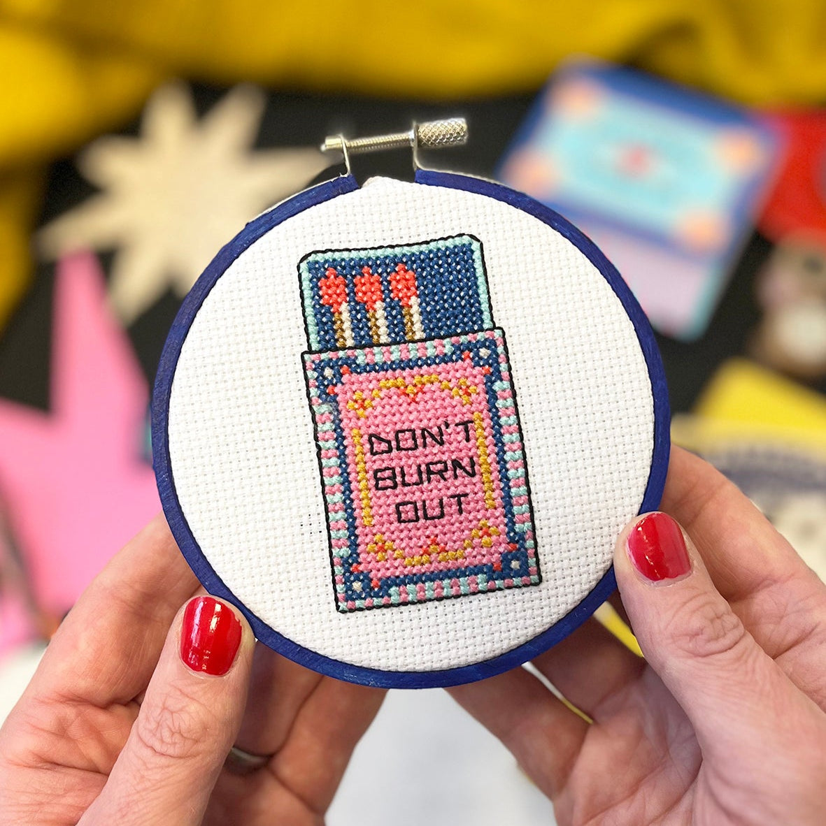 Cross-stitched hoop with 'Don't Burn Out' design held by a person with red nail polish.