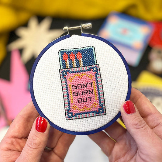 Cross-stitched hoop with 'Don't Burn Out' design held by a person with red nail polish.