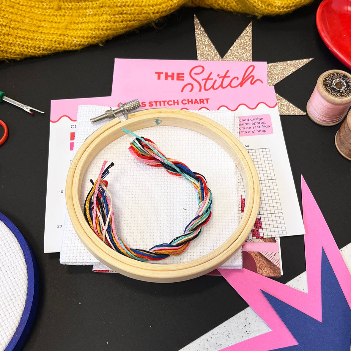 Embroidery hoop with colorful thread on a black surface with 'The Stitch' chart and yarn.