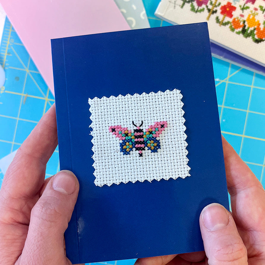 Blue book with a cross-stitched butterfly design held by hands on a blue cutting mat.