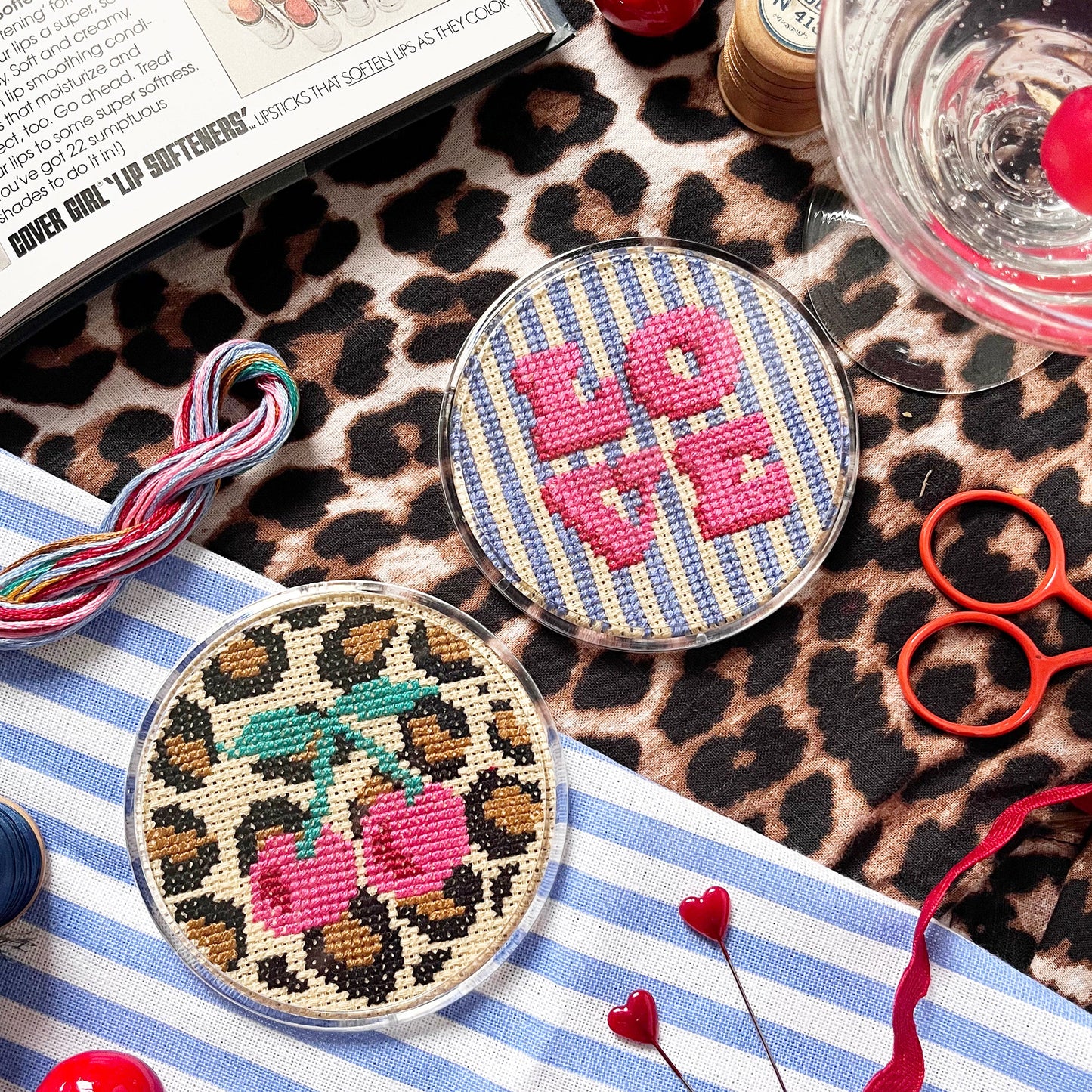 Two cross stitched coasters on a striped fabric with sewing supplies.