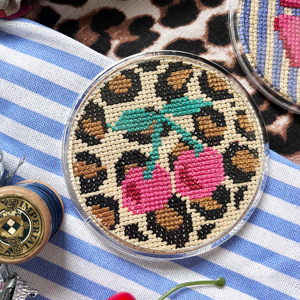 Round cross stitched coaster with leopard print and cherry design on a striped surface.
