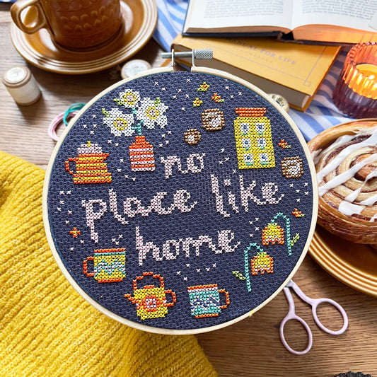 Embroidery hoop with 'No place like home' text and cross-stitch design on a table with coffee and books.