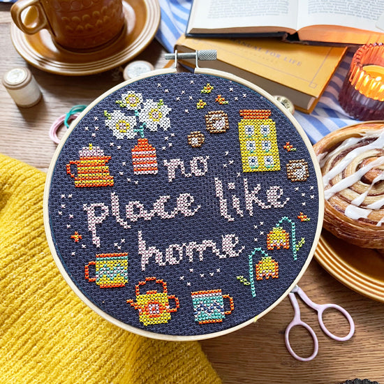 Embroidery hoop with 'No place like home' text and cross-stitch design on a table with coffee and books.