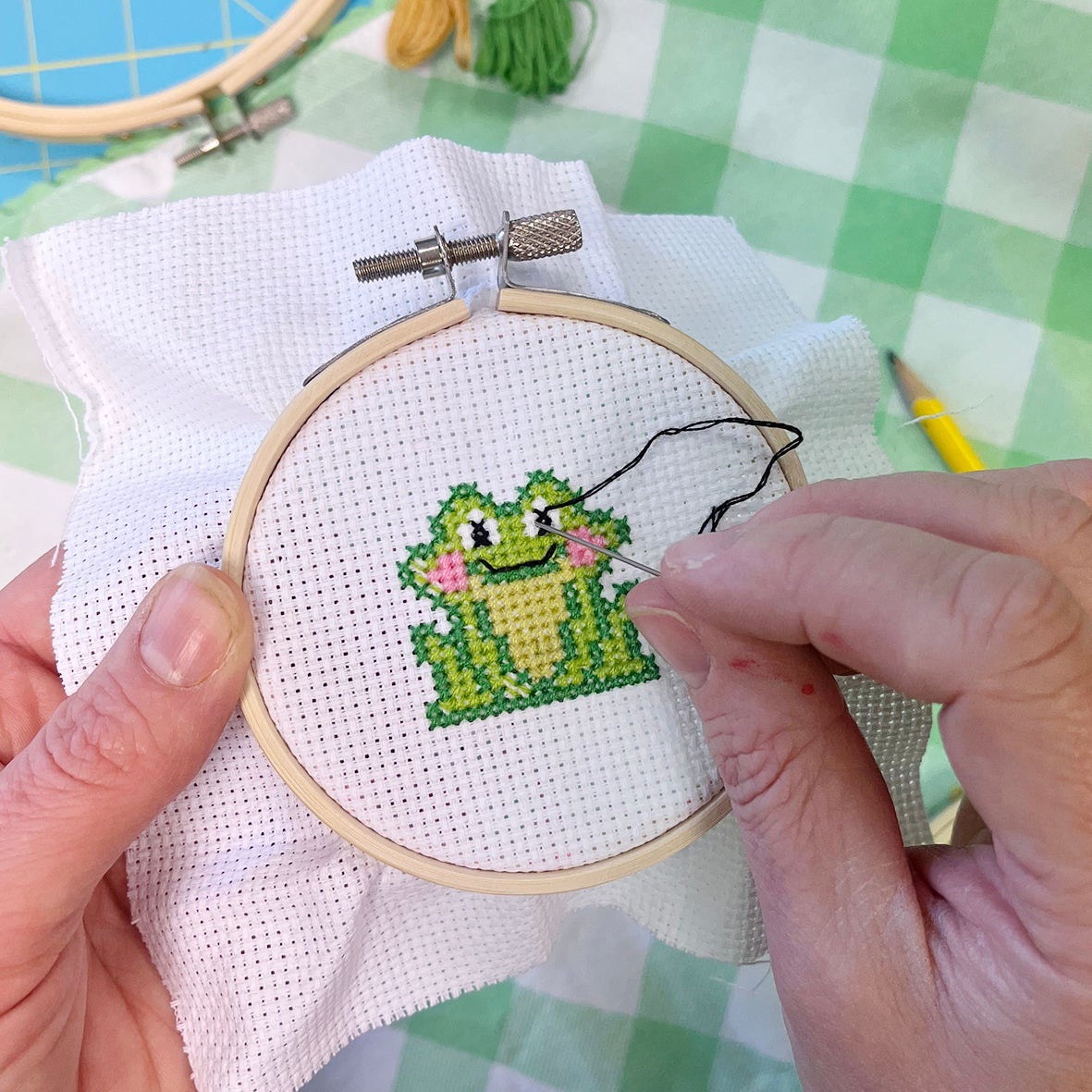 Cross-stitching a green frog design on fabric with an embroidery hoop.